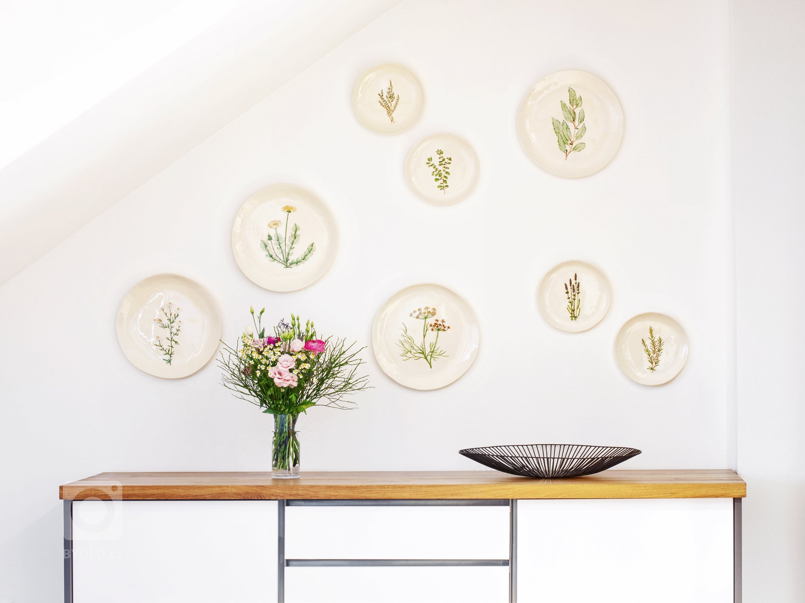 Stěnu dekorují keramické talíře s ruční kresbou bylinek podle starých herbářů. Prostřený stůl zdobí lněné prostírání šité na míru.