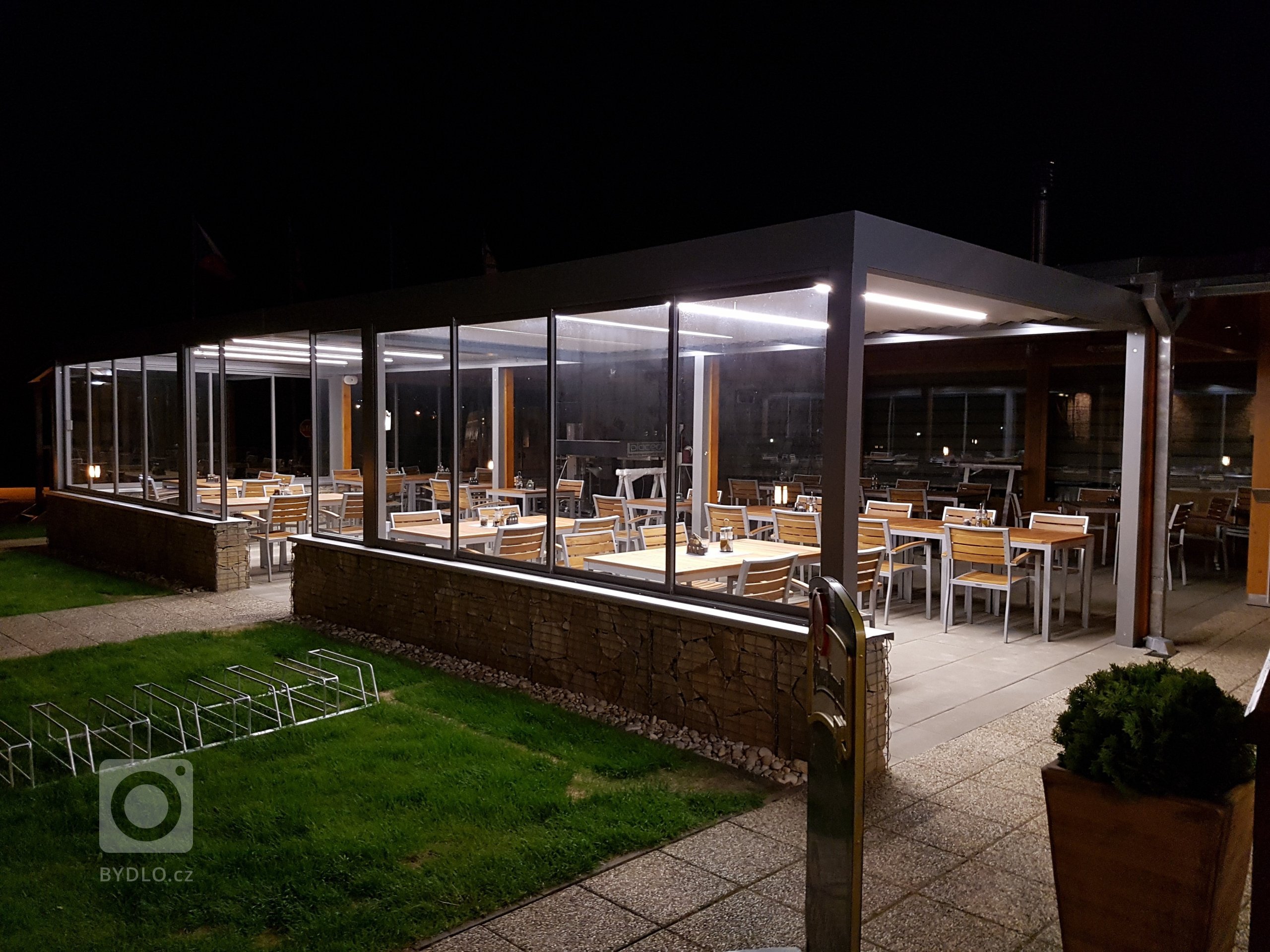 Hliníkovú pergolu jsme osvětlili a zavedli do ní všechny inženýrské sítě, díky čemuž se stala plnohodnotnou venkovní restaurací a barem.&nbsp;Boční zakrytí…