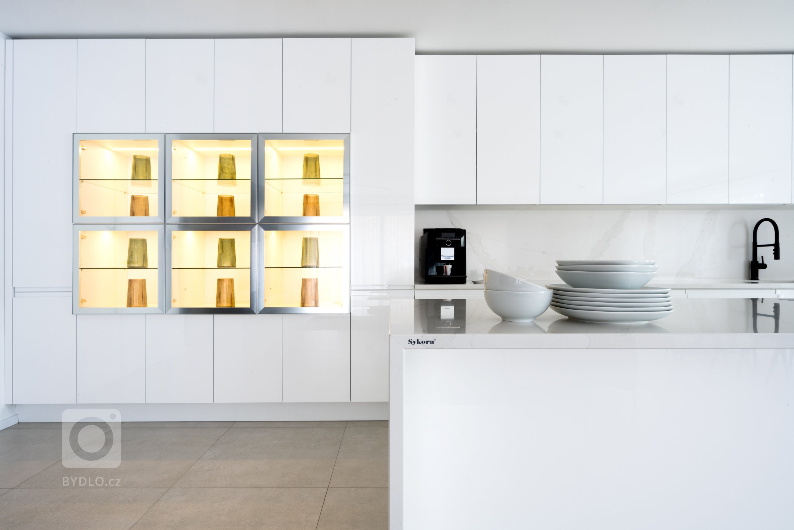 Specialitou sestavy, něčím, co originálně vybočuje z šablon, je prosklená vitrína v levé části vysokých skříní. Prosvětluje už tak jasný prostor, a díky vnitřnímu podsvícení mu dodává až magickou hladivost.